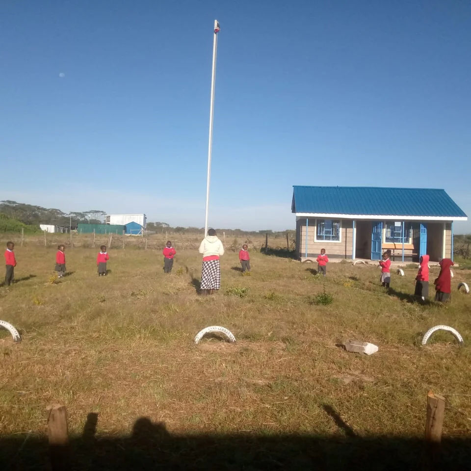 Learners and an adult gathered around the flagpole outside a Namunyak school building